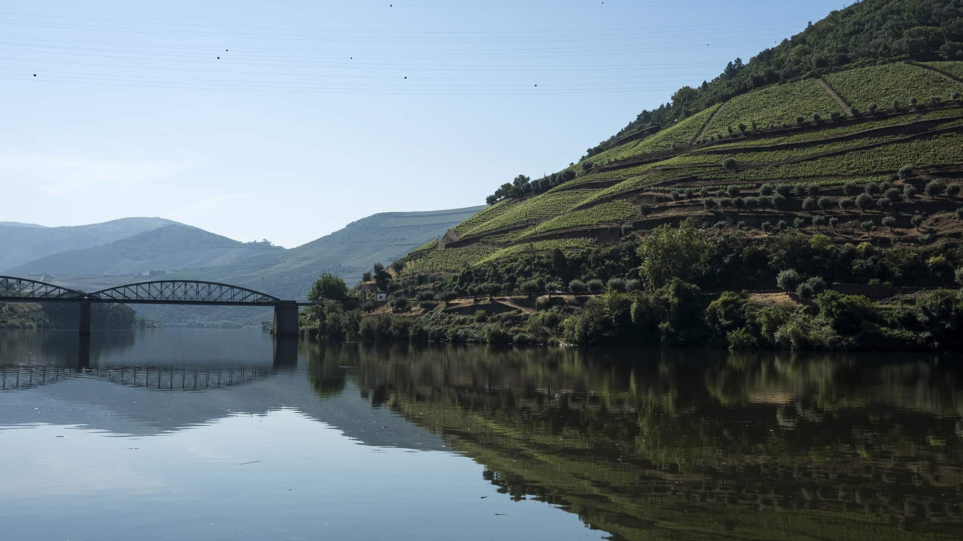 A river reflects terraced green hills and a metal bridge under a clear sky, with mountains in the background.