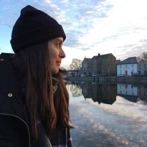A woman in a black beanie and coat stands by a calm river, with buildings and trees reflected in the water under a cloudy sky.