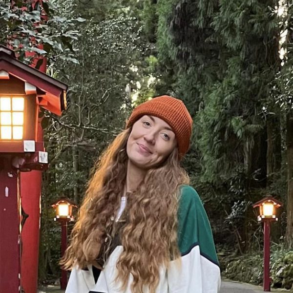 A person with long wavy hair and an orange beanie stands outdoors by red lanterns and tall trees, smiling at the camera.