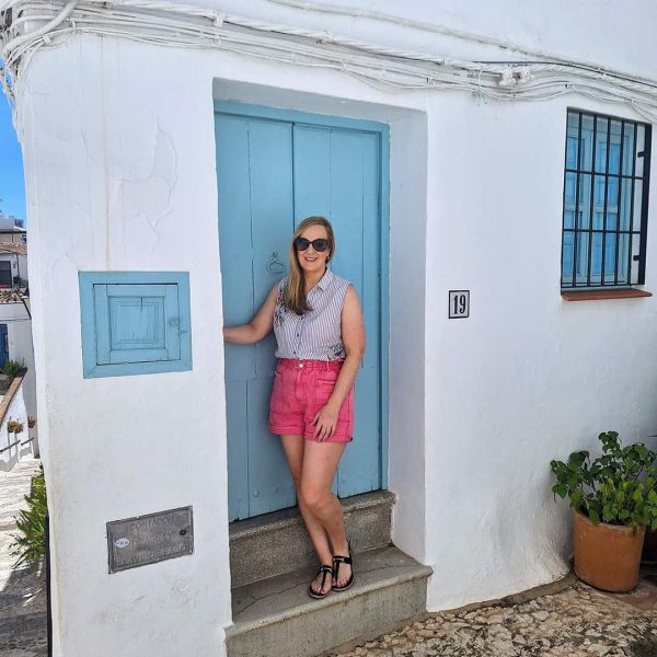 A woman wearing sunglasses, a sleeveless top, and pink shorts stands in front of a blue door on a white building, numbered 19, with potted plants nearby.