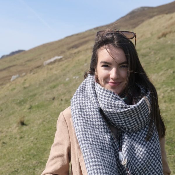 A woman with long brown hair wearing a large houndstooth scarf and a beige coat stands outside on a grassy hill with blue sky in the background.