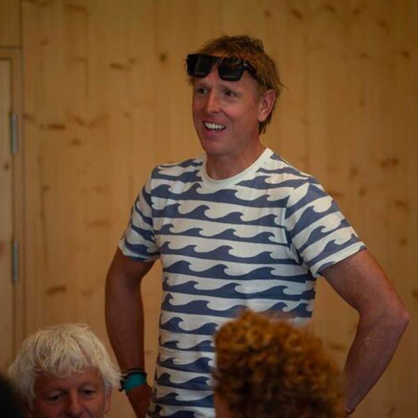 A man wearing a white and blue wave-patterned shirt and sunglasses on his head stands with hands on hips indoors, with others seated nearby.