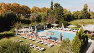 Outdoor swimming pool surrounded by sun loungers and umbrellas, with trees and a stone wall in the background on a sunny day.