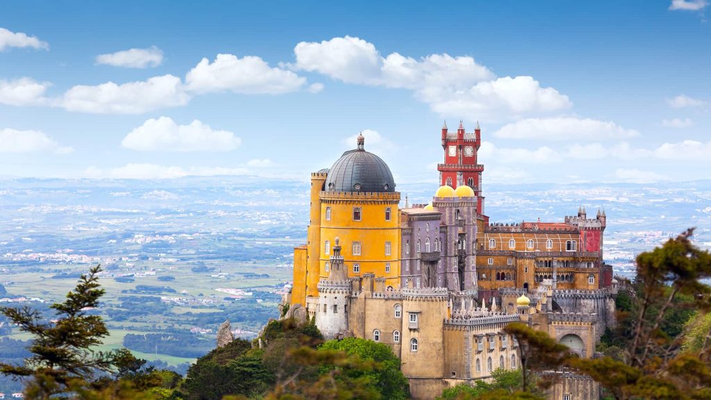 Colorful hilltop castle with yellow and red towers, set against a panoramic landscape and blue sky with scattered clouds.