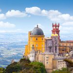 Colorful hilltop castle with yellow and red towers, set against a panoramic landscape and blue sky with scattered clouds.