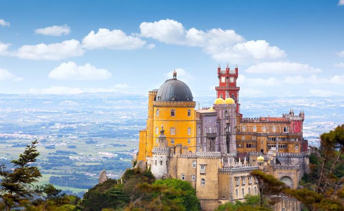 Colorful hilltop castle with yellow and red towers, set against a panoramic landscape and blue sky with scattered clouds.