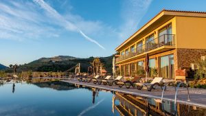 Modern hotel building with large windows and balconies beside an outdoor pool with lounge chairs, surrounded by landscaped hills under a clear blue sky.