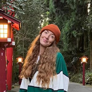 A person with long curly hair wearing a red beanie and green striped jacket stands on a path beside red lanterns and tall trees. A person with long curly hair wearing a red beanie and green striped jacket stands on a path beside red lanterns and tall trees.