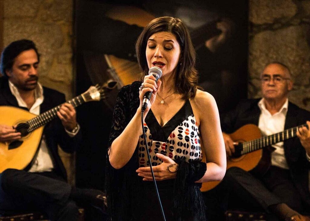 A woman sings into a microphone while two men seated behind her play acoustic guitars, creating a lively atmosphere reminiscent of small group travel experiences in Portugal.