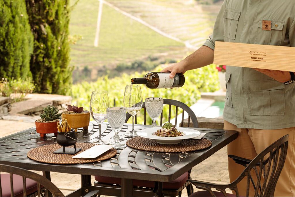 A person pours white wine into glasses on an outdoor table set with plates of food and decorative touches, as a small group travel experience unfolds amid vineyard scenery in the heart of Portugal.