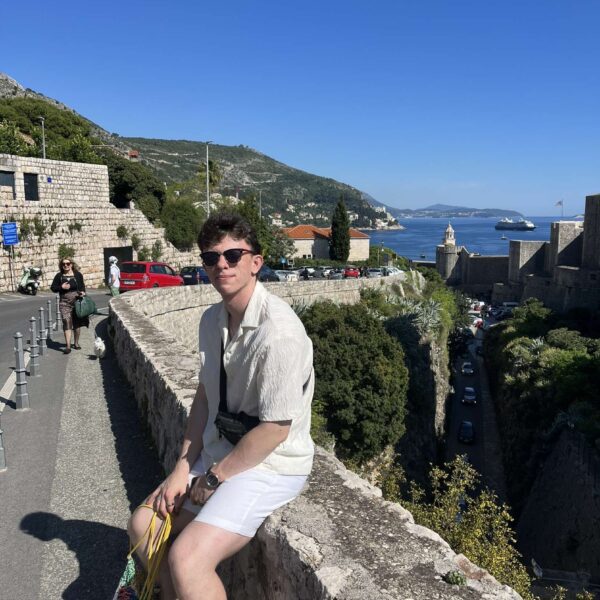 A man in sunglasses and summer clothes sits on a stone wall near a road, with greenery, hills, and the sea in the background on a sunny day.