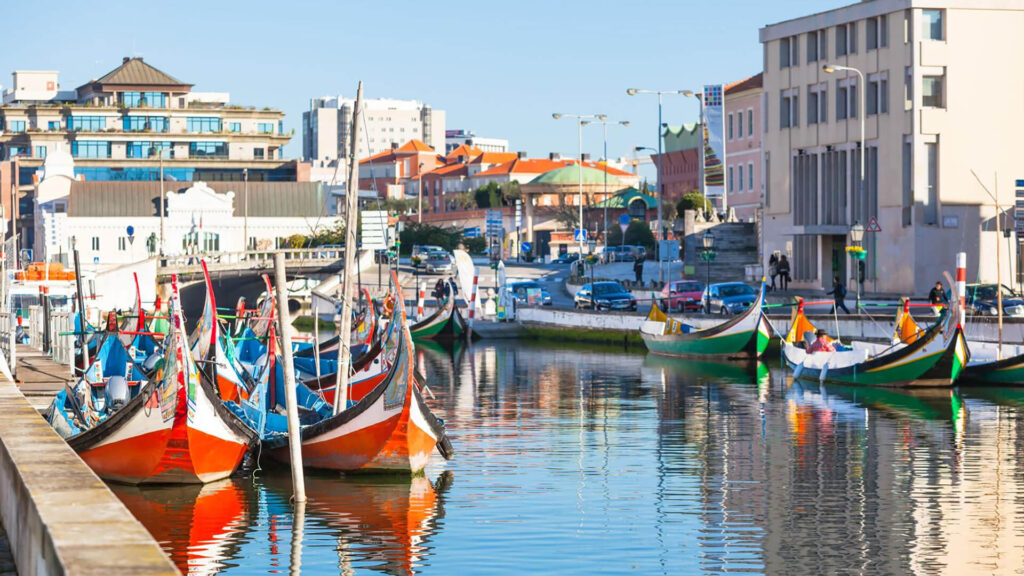 Colorful gondola-style boats are docked along a canal lined with buildings and cars under a clear blue sky in a cityscape.