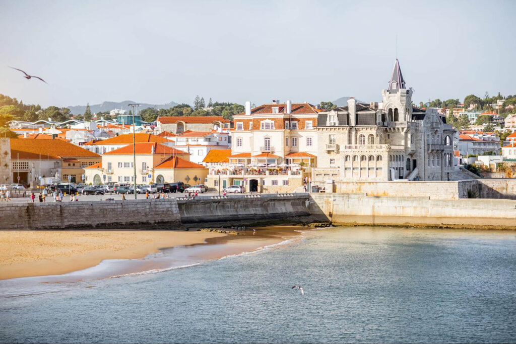A seaside town with red-roofed buildings, a historic stone mansion, a sandy beach, and people walking along a waterfront promenade.