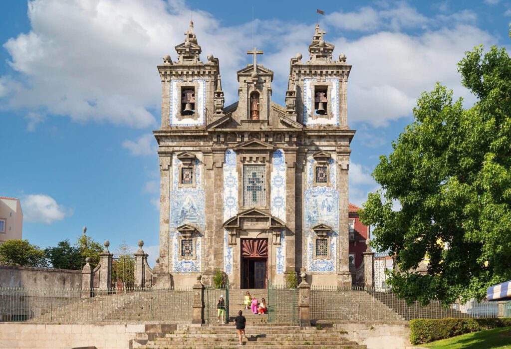 Stone church with blue and white tile facade, two bell towers, and a large staircase in front; several people are visible on the steps. A must-see when you visit Portugal, especially during spring as suggested by the Portugal seasonal guide.