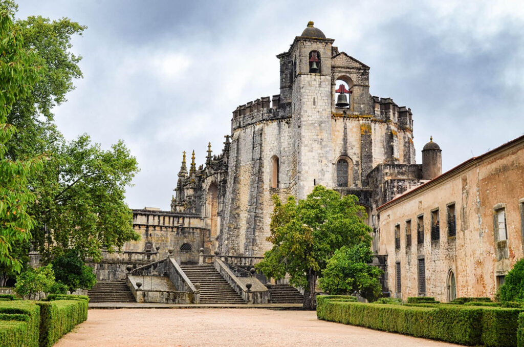 Stone castle-like building with a large staircase, arched windows, and a bell tower, surrounded by trees and greenery under a cloudy sky.