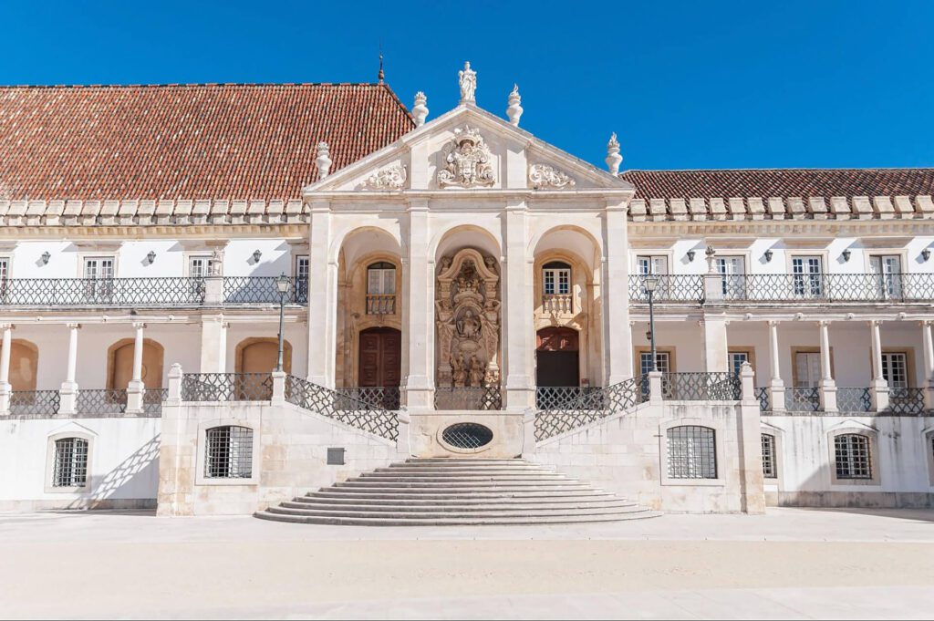 Historic building with a central staircase leading to an ornate entrance, flanked by columns and sculptures, with a tiled roof and blue sky above.