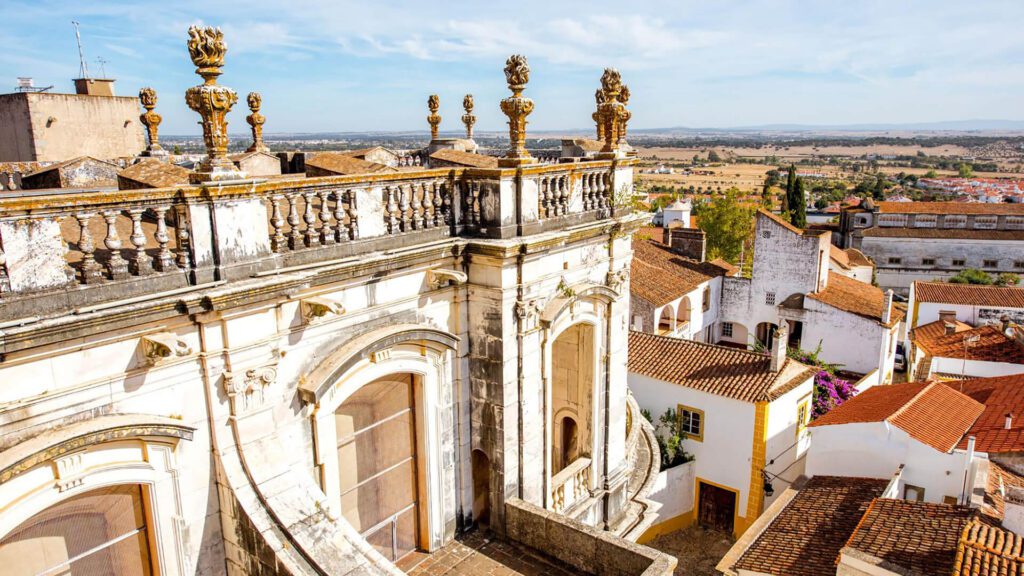 Ornate stone balcony with decorative columns overlooks red-roofed buildings and a rural landscape under a blue sky.