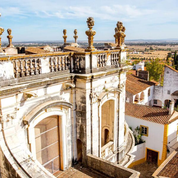Ornate stone balcony with decorative columns overlooks red-roofed buildings and a rural landscape under a blue sky.