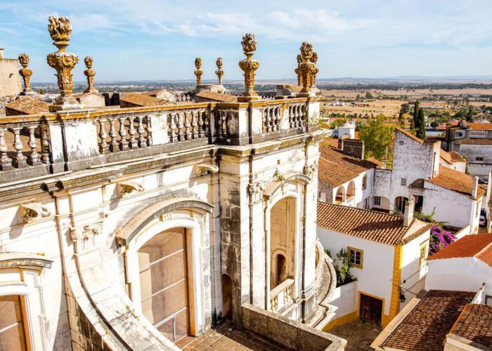 Ornate stone balcony with decorative columns overlooks red-roofed buildings and a rural landscape under a blue sky.
