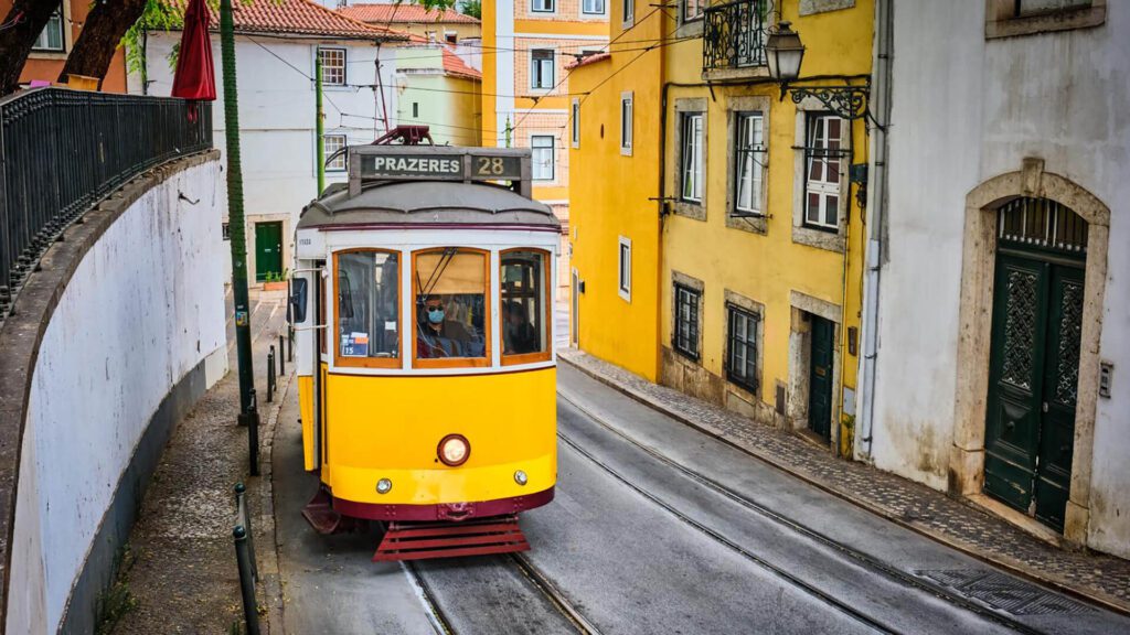 A yellow tram labeled "28 Prazeres" travels along tracks between colorful buildings on a narrow street in Lisbon, Portugal—a classic scene for any First-Time Visitor seeking authentic Portugal Travel Tips.