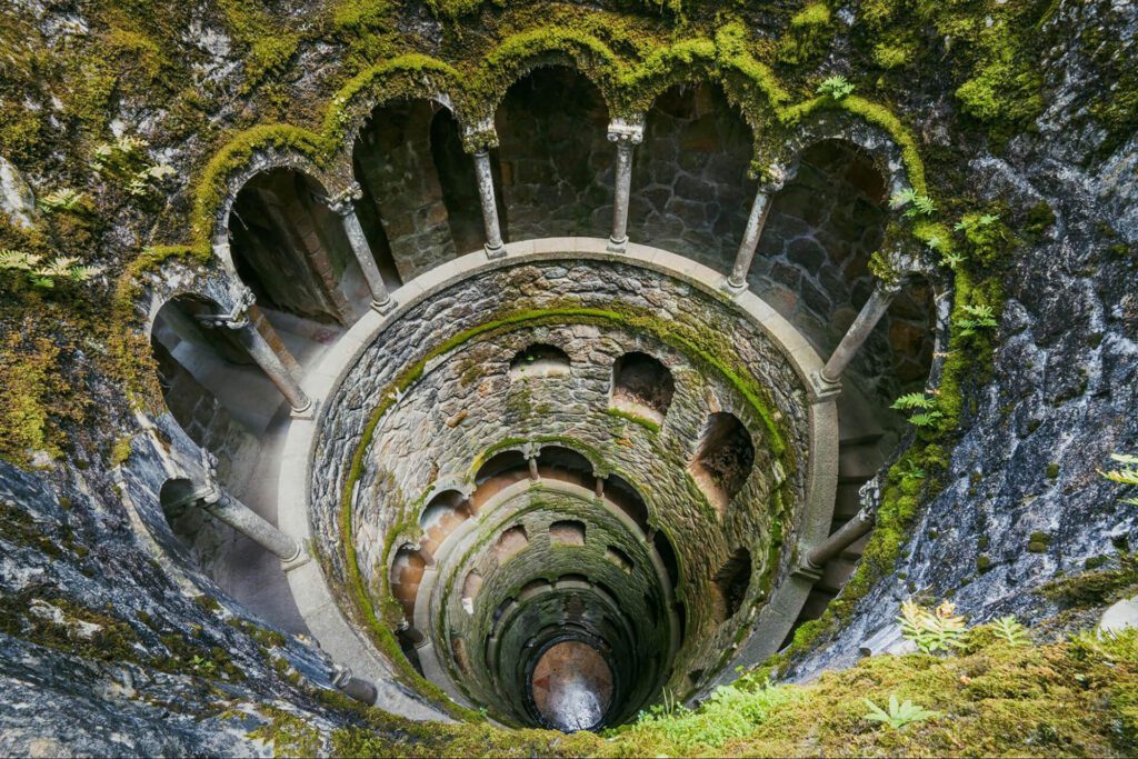 A stone spiral staircase descends into a deep, circular well with moss-covered walls and arched openings at each level.