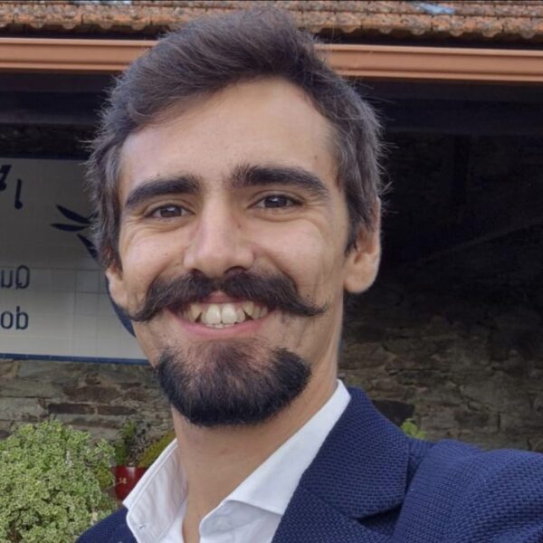 A man with brown hair, a styled mustache, and a beard is smiling at the camera, wearing a navy blazer and white shirt, with a building and greenery in the background.