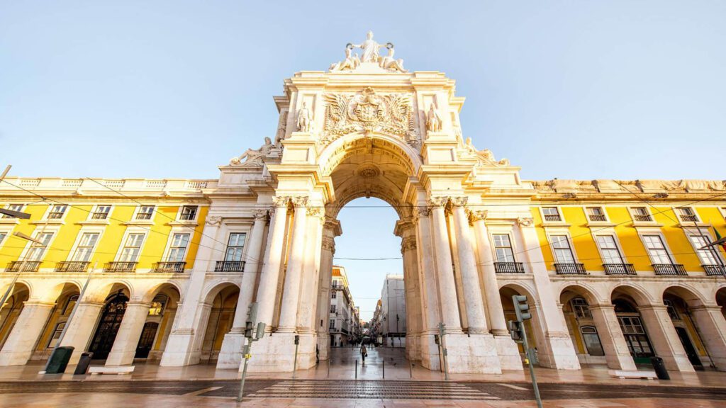 The image shows the Arco da Rua Augusta, a large stone triumphal arch with sculptures, flanked by yellow buildings in Lisbon, Portugal, under a clear sky.