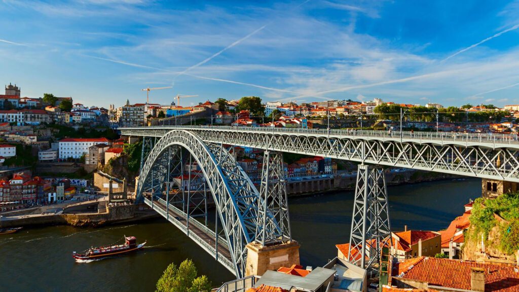 Large iron arched bridge spans a river with colorful buildings and a boat below, under a blue sky with scattered clouds and contrails.