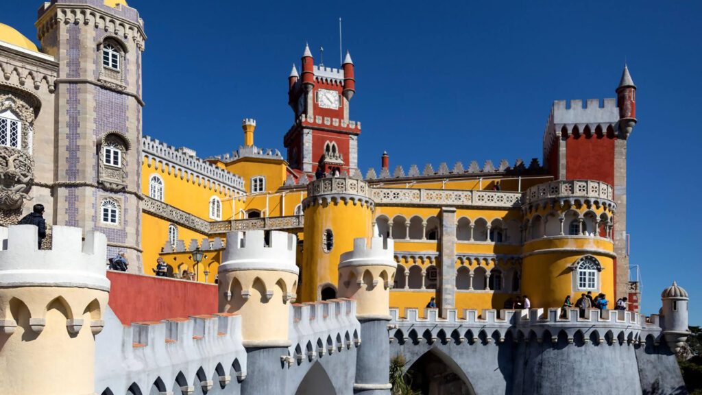 The image shows the colorful, detailed exterior of Pena Palace in Sintra, a highlight for Portugal tourism, featuring yellow, red, and grey towers set against a clear blue sky.