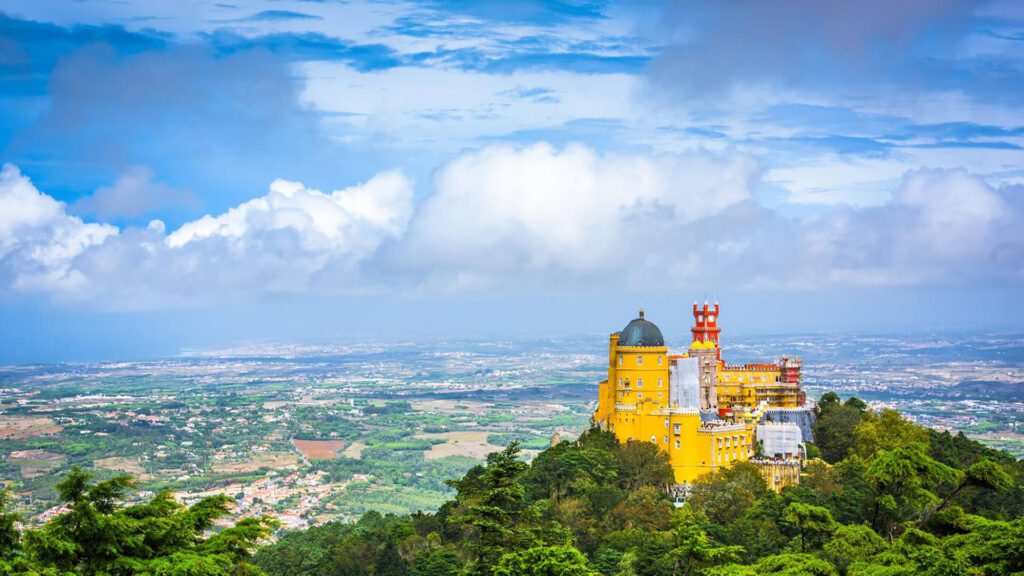 A colorful, hilltop palace surrounded by green forest overlooks a vast, flat landscape under a partly cloudy sky.