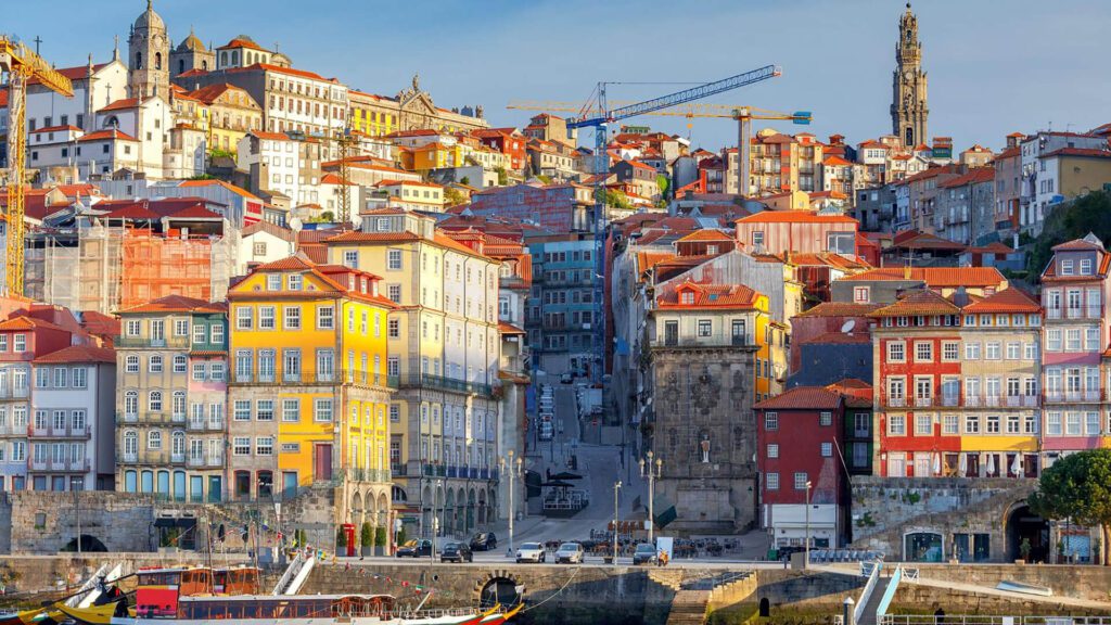 Colorful hillside buildings in Porto, Portugal, rise above a lively waterfront dotted with boats—one of the top Portugal attractions and a highlight for anyone seeking the Best Things to Do in Portugal. Construction cranes and a church tower complete the scenic view.