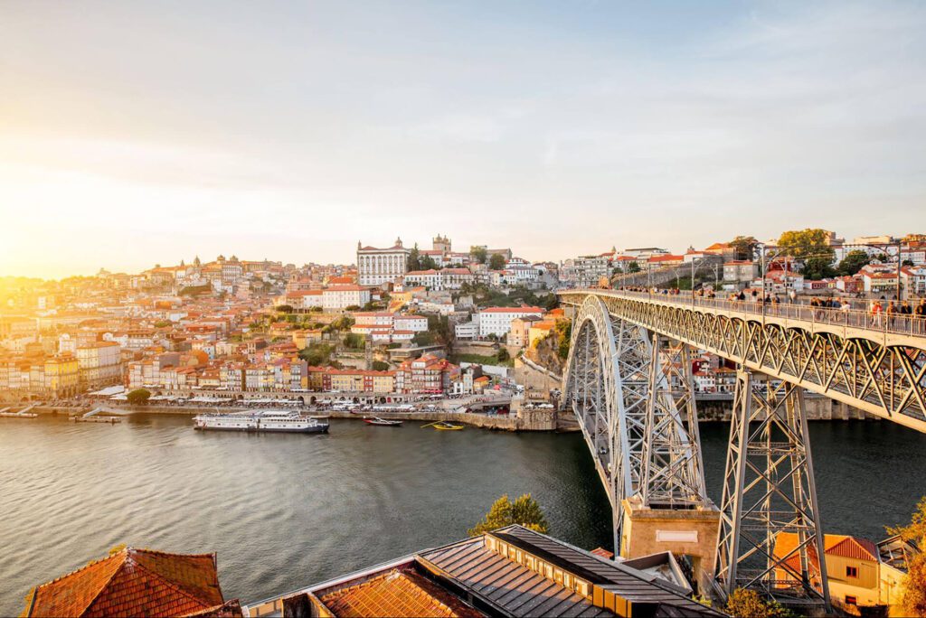 A large metal bridge spans a river, with a city of colorful buildings and hillsides in the background under a clear sky—a picture-perfect spot highlighted in any Portugal seasonal guide if you’re planning to visit Portugal.
