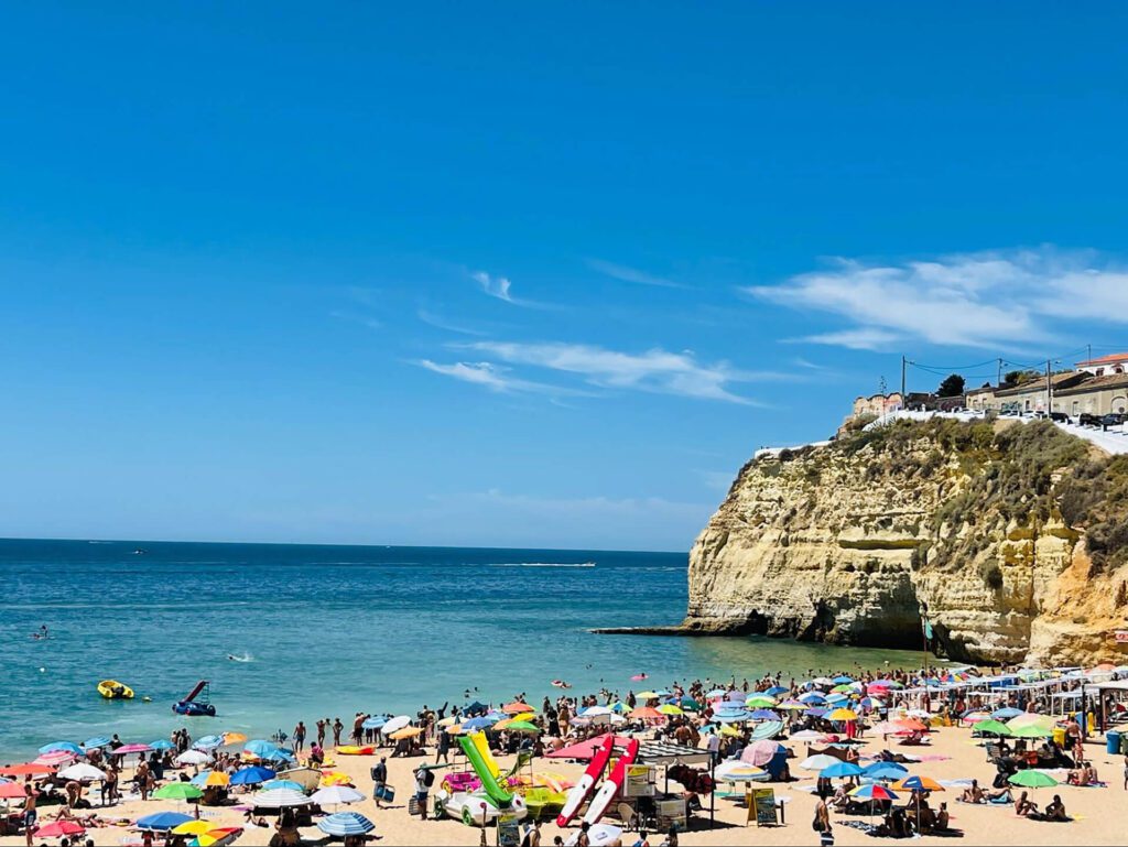 Crowded beach with colorful umbrellas and people by the sea; rocky cliffs and buildings overlook the shoreline under a clear blue sky, capturing summer scenes found in any Portugal travel guide.