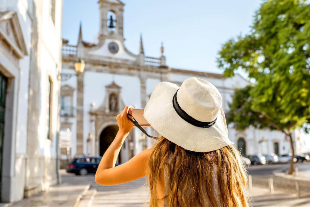 A woman in a white sunhat takes a photo of a historic building with a bell tower on a sunny day, capturing memories as a first-time visitor to Portugal.