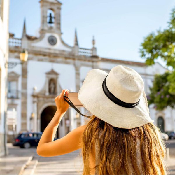 A woman in a white sunhat takes a photo of a historic building with a bell tower on a sunny day, capturing memories as a first-time visitor to Portugal.