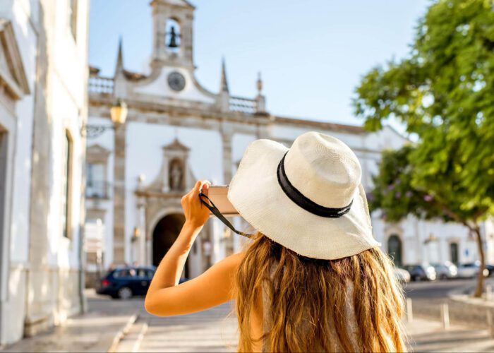 A woman in a white sunhat takes a photo of a historic building with a bell tower on a sunny day, capturing memories as a first-time visitor to Portugal.