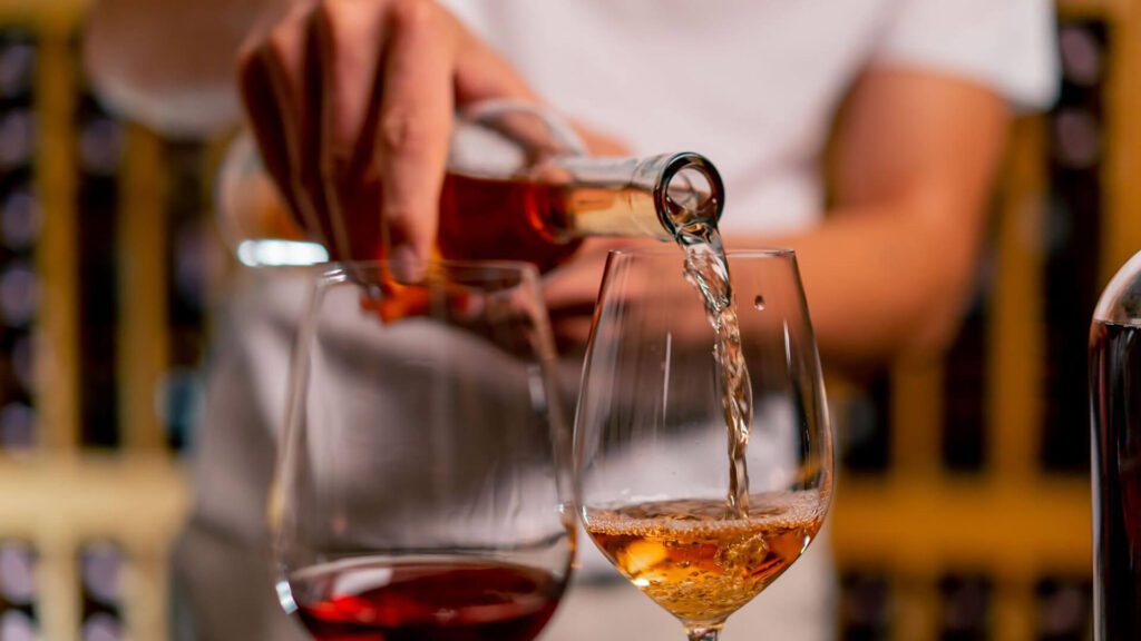 A person pours white wine into a glass beside a glass of red wine on a table, with wine racks blurred in the background—capturing the vibrant wine culture among the best things to do in Portugal.