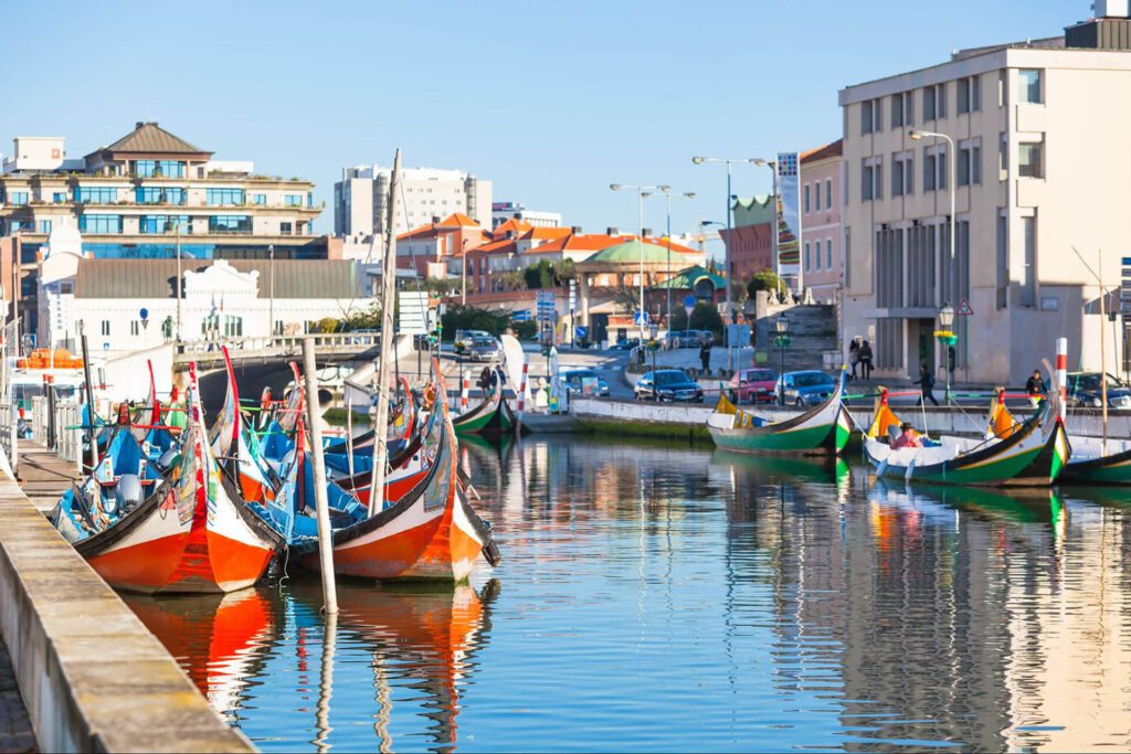 Several colourful boats are moored along a canal lined with buildings in a sunny urban area, perfect for featuring on your Portugal itinerary.