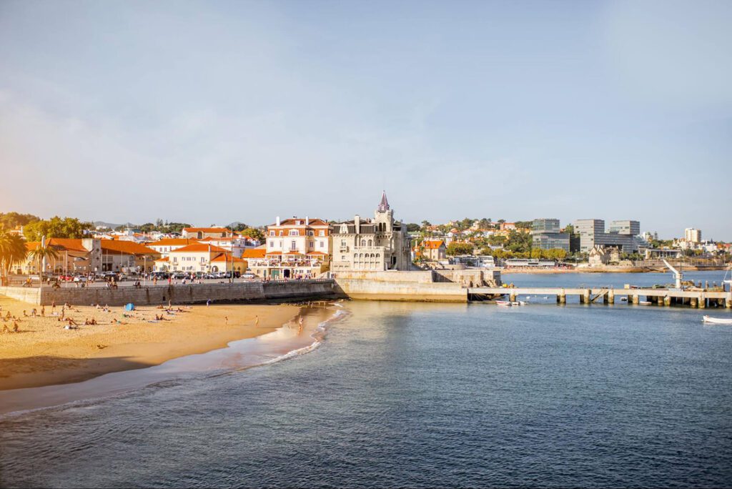 View of a coastal town with a sandy beach, historic building, and pier extending into the calm sea; people are relaxing on the beach and modern buildings are visible in the background—a perfect stop on your best Portugal holiday or 7-day itinerary.