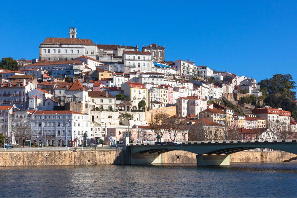 A view of Coimbra, Portugal, showing historic white and pastel buildings on a hillside above the Mondego River with a bridge in the foreground under a clear blue sky—a must-see stop on any 7-Day Itinerary Portugal adventure.