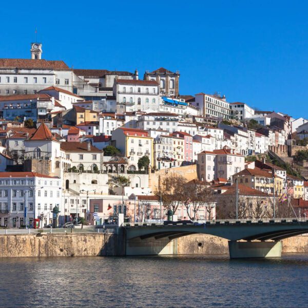 A view of Coimbra, Portugal, showing historic white and pastel buildings on a hillside above the Mondego River with a bridge in the foreground under a clear blue sky—a must-see stop on any 7-Day Itinerary Portugal adventure.