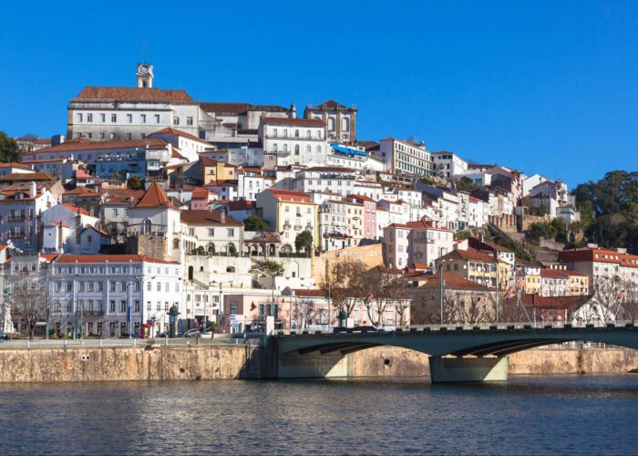 A view of Coimbra, Portugal, showing historic white and pastel buildings on a hillside above the Mondego River with a bridge in the foreground under a clear blue sky—a must-see stop on any 7-Day Itinerary Portugal adventure.