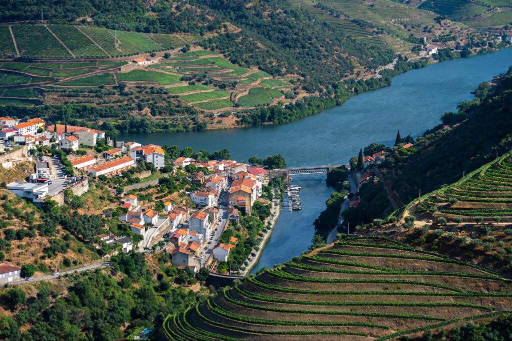 Aerial view of a riverside village with red-roofed buildings, surrounded by terraced vineyards and green hills—a must-see stop on any best Portugal itinerary.