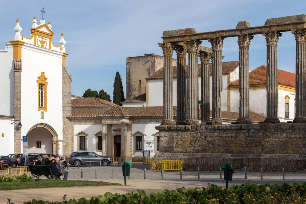 A historic Roman temple of Corinthian columns stands beside a white and yellow church in a lively town square, a must-see on any best Portugal itinerary, with people sitting on benches and soaking up the atmosphere.