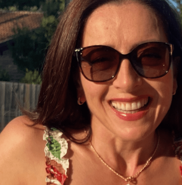 A woman wearing sunglasses and a floral dress smiles outdoors on a sunny day, with trees and a house in the background.