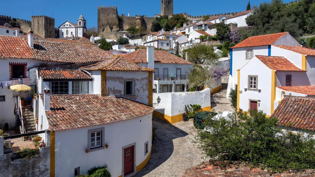 Discover one of the underrated places in Portugal: a historic village with whitewashed houses, red tiled roofs, cobbled streets, and a hilltop castle beneath a clear blue sky.