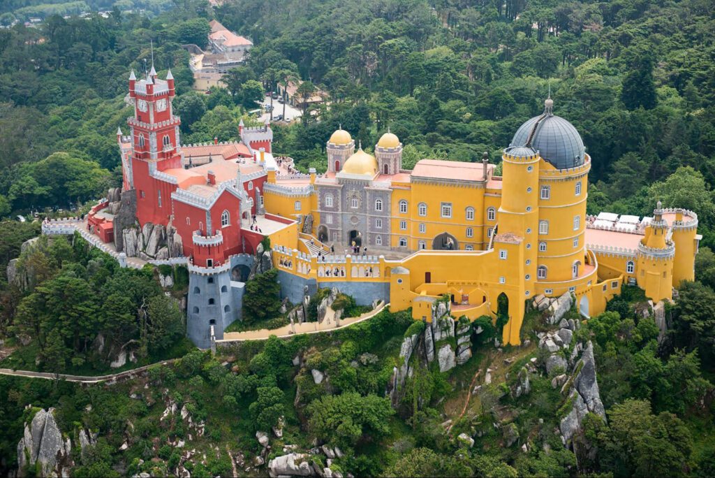 Aerial view of Pena Palace in Sintra, Portugal, featuring bright red and yellow buildings with towers and domes, surrounded by green forested hills—a must-see on any best Portugal itinerary.