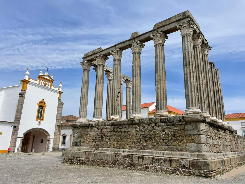 Ancient stone columns of the Roman Temple of Évora stand beside a white church under a blue sky with streaks of cloud, making it one of the most underrated places in Portugal and truly Portugal worth visiting.