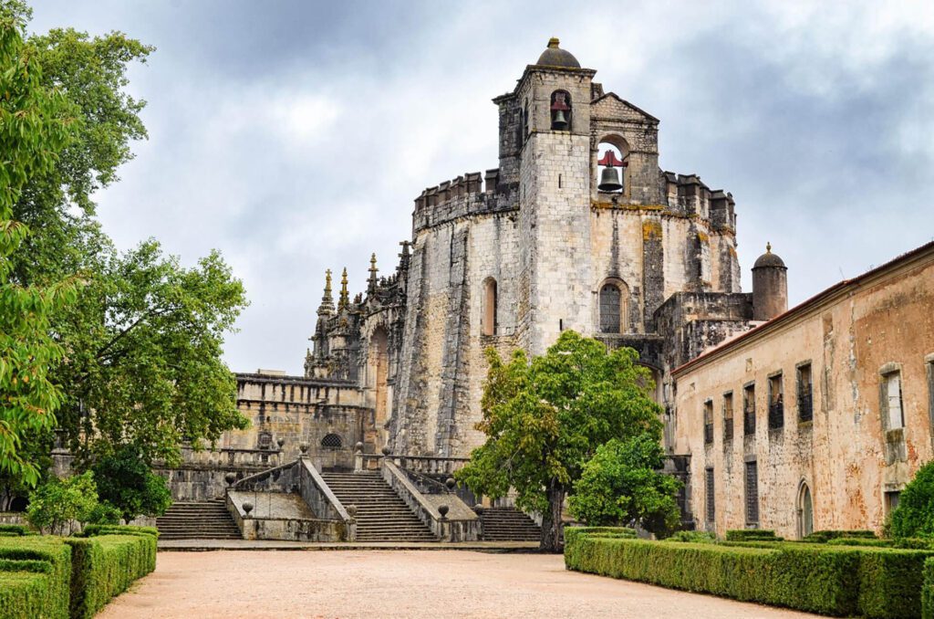 This stone medieval castle, with its round tower, arched windows, and bell, surrounded by trees and hedges under a cloudy sky, is one of the underrated places in Portugal worth visiting.