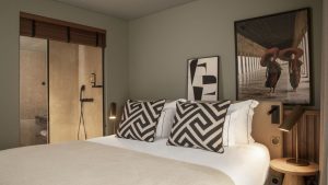 A neatly made bed with patterned cushions, a beige blanket, modern wall art, and a glass-walled bathroom in a contemporary hotel room—perfect for relaxing after your 8-Day Portugal Tour.